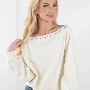 A blonde woman wearing a cream-colored, long-sleeve off-the-shoulder top with silver stud detailing along the neckline, paired with blue jeans, against a plain white background.